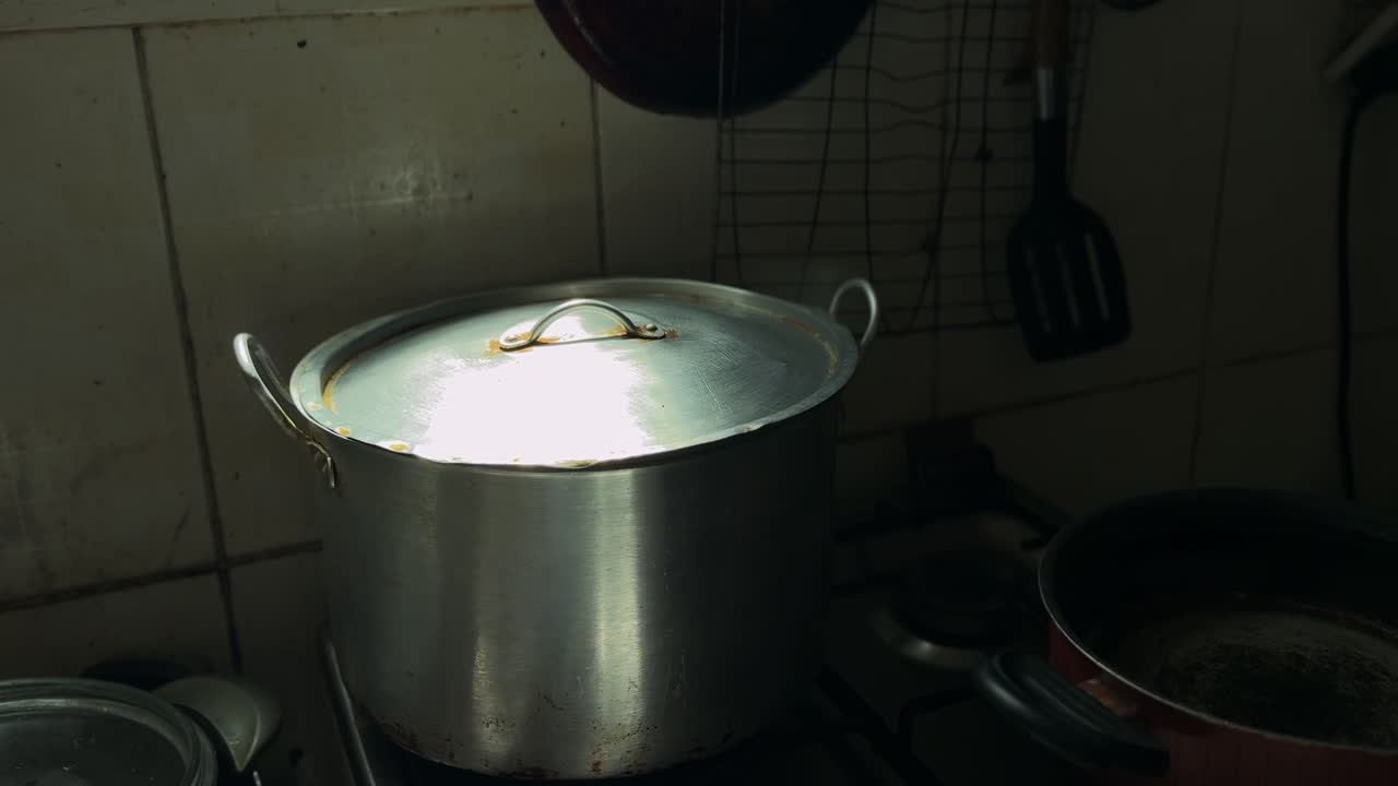 En stor kokegryte av metall med lokk som varmes på en gasskomfyr i et dunkelt, rustikt kjøkken, med damp, kjøkkenutstyr og stemningsfullt innelys som understreker hjemlig matlaging.