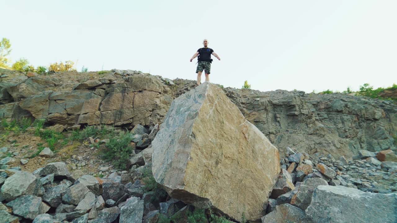 a brave traveler is standing on top of a huge rock with his hands raised on the background of a beautiful location in the summer