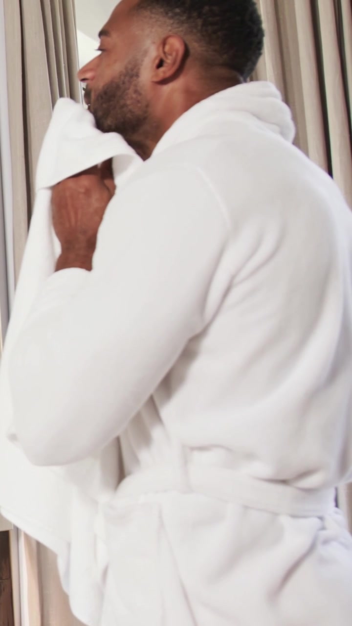Vertical video: Holding towel to face, man in bathrobe drying and checking jawline near curtains
