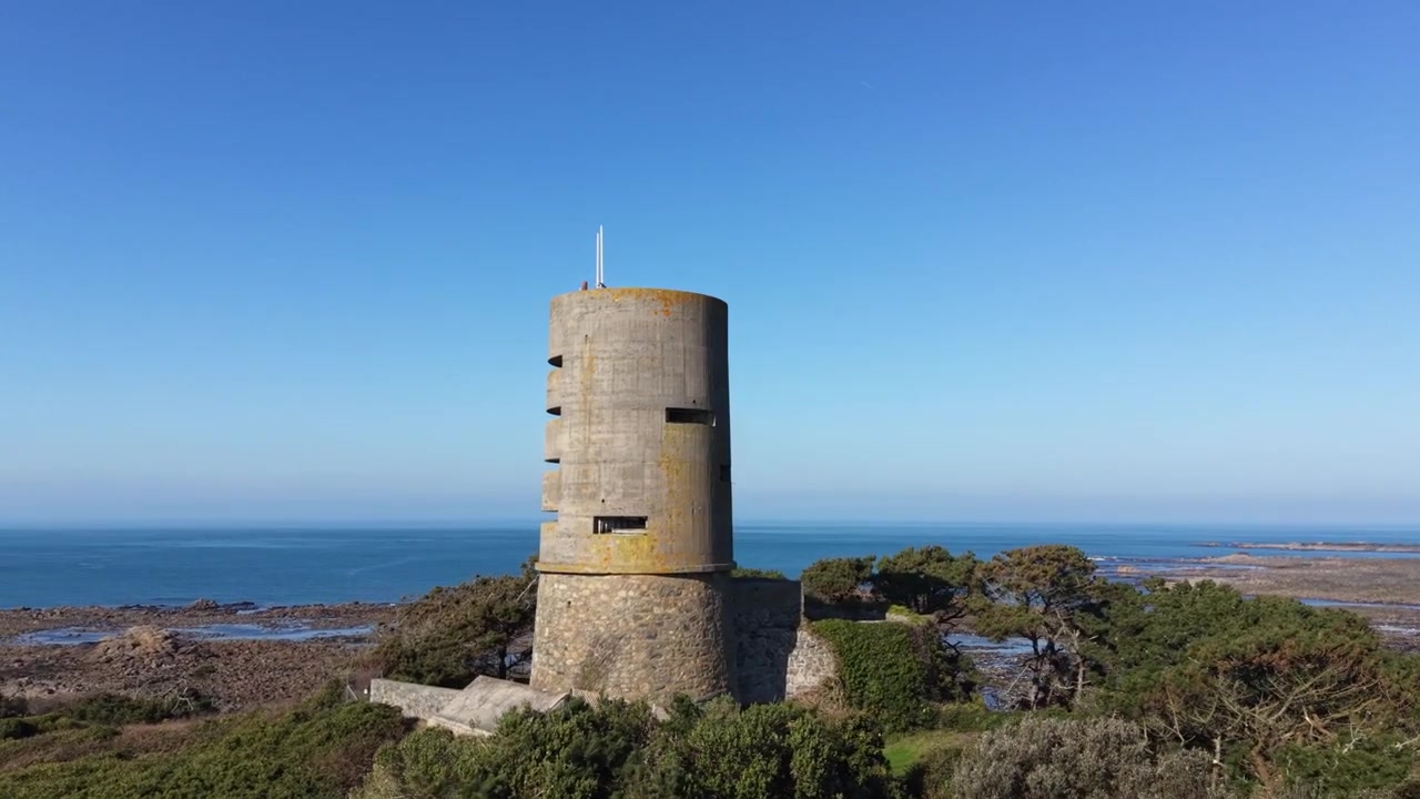Imágenes de dron en círculo cercano de la antigua torre de vigilancia alemana en el promontorio de L'Eree, Guernsey, con vistas al mar y a la isla de Lihou, bajo el sol de la tarde y un impresionante cielo azul claro.