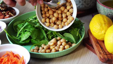 Creating-a-salad-adding-chick-peas-to-plate-with-spinach-and-carrots-slices-cilantro-onions-sliced-almonds-dressing-lemons