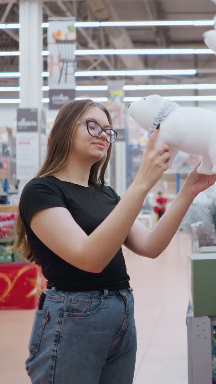 una mujer joven con gafas y camisa negra examina pensativamente un juguete blanco de peluche de oso polar en una tienda de vacaciones, con otro cliente pasando en el fondo en medio de decoraciones festivas