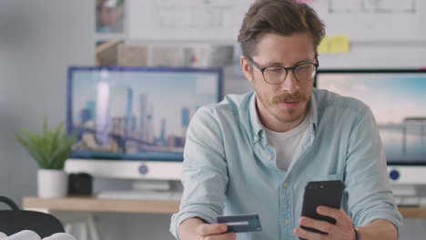 Arquitecto-Masculino-En-La-Oficina-Trabajando-En-El-Escritorio-Haciendo-Compras-En-Línea-Usando-Tarjeta-De-Crédito-En-El-Teléfono-Móvil