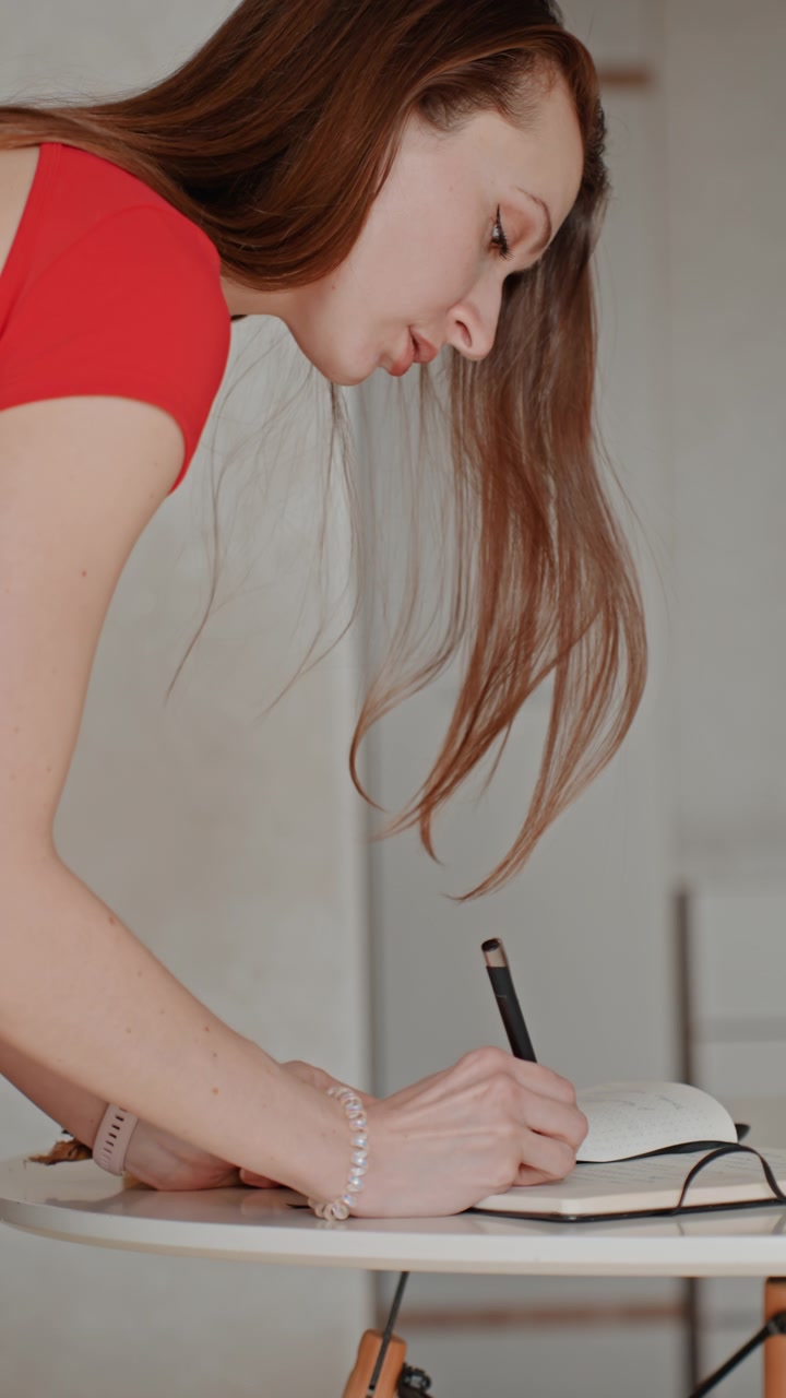 Giovane donna caucasica che scrive al tavolo di casa, sfogliando le pagine di un quaderno, prendendo appunti con una penna nera, occhiali e un biscotto accanto al quaderno, chinata sulla superficie in un ambiente di cucina moderna, mattina mite.