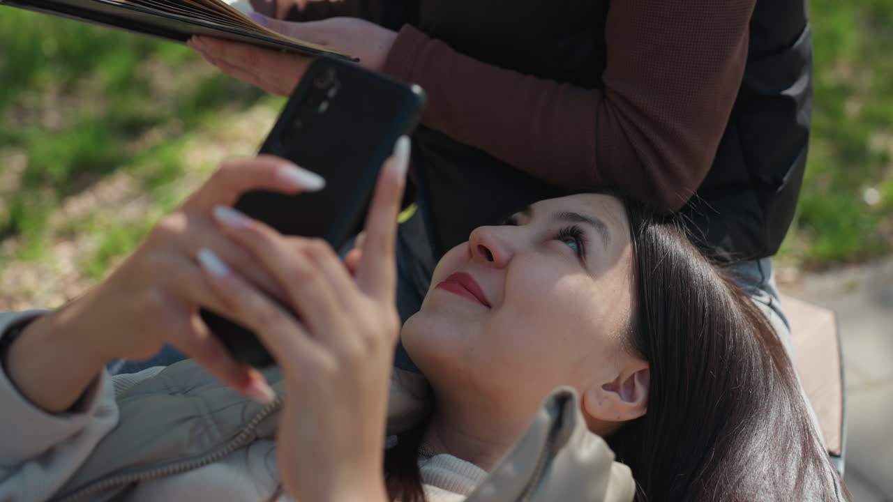 흰색 배경의 여성이 피크닉에서 소설을 소리 내어 읽고, 아시아인 친구는 담요와 펼쳐진 페이지가 있는 봄날의 문학 클럽 분위기 속에서 휴대폰을 확인하며 듣고 있습니다. 따뜻하고 솔직한 상호 작용과 사려 깊은 토론이 느껴집니다.