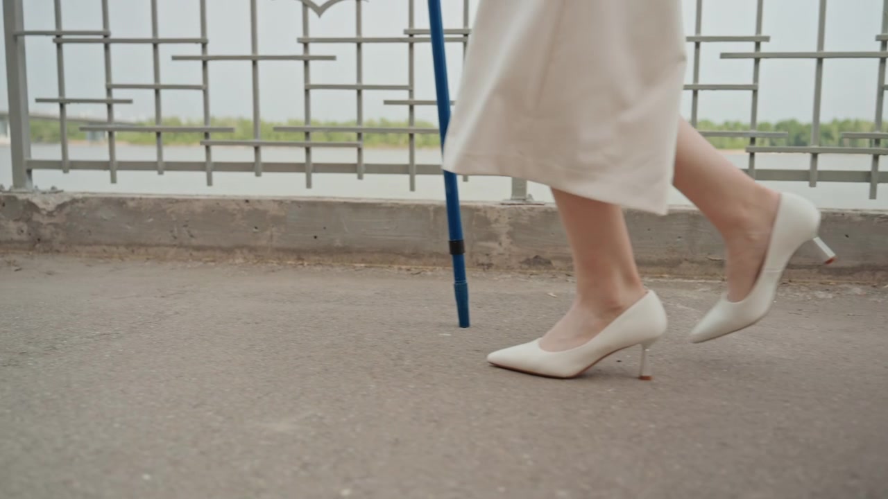 Una donna in abito bianco cammina lungo un sentiero all'aperto, tenendo e usando un bastone blu per supporto. La scena si concentra sulla parte inferiore del suo corpo e sul movimento attraverso un ambiente urbano che include elementi metallici.