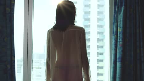 woman in a shear shirt with the light silhouetting her perfect body opens the bedroom curtains to let in the light and greet a new day of opportunities