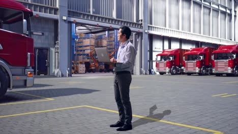 full body side view of an asian male professional worker, outside of logistics distributions warehouse, observes by looking up before he come to concentrating on the laptop and keep on typing