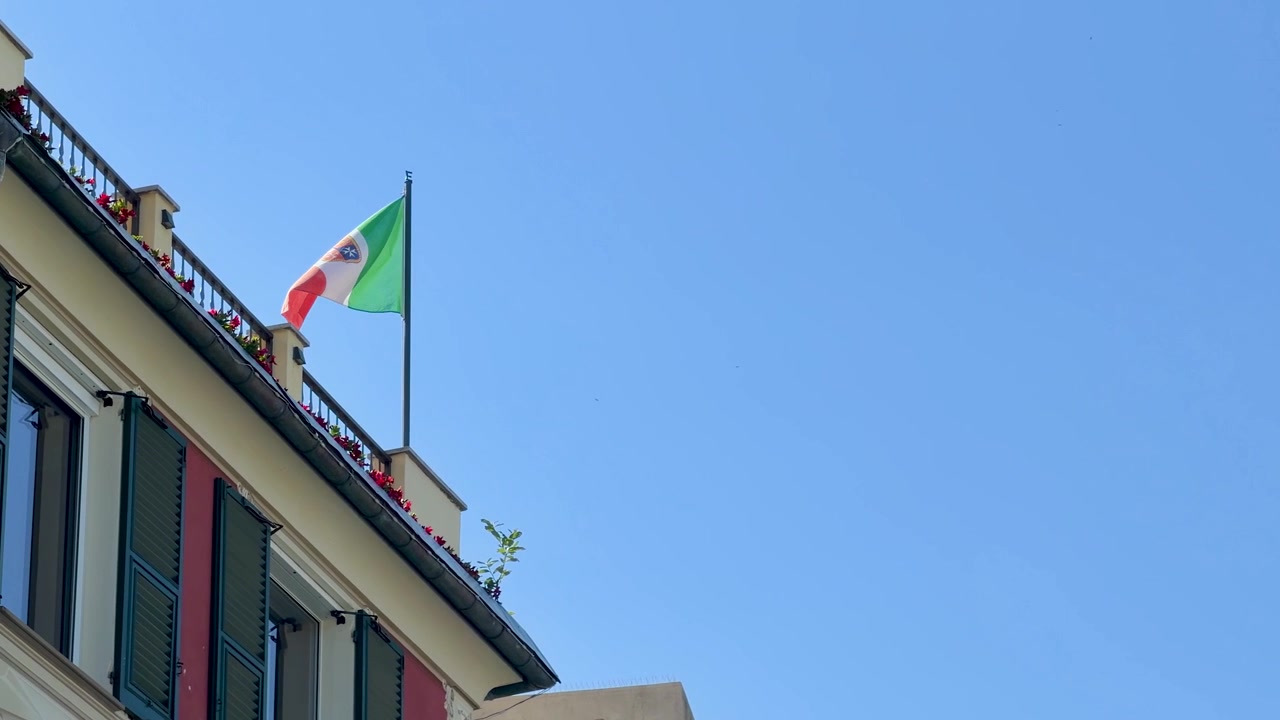 Inquadratura dal basso di una bandiera nazionale che sventola da un balcone mediterraneo sotto un cielo blu.
