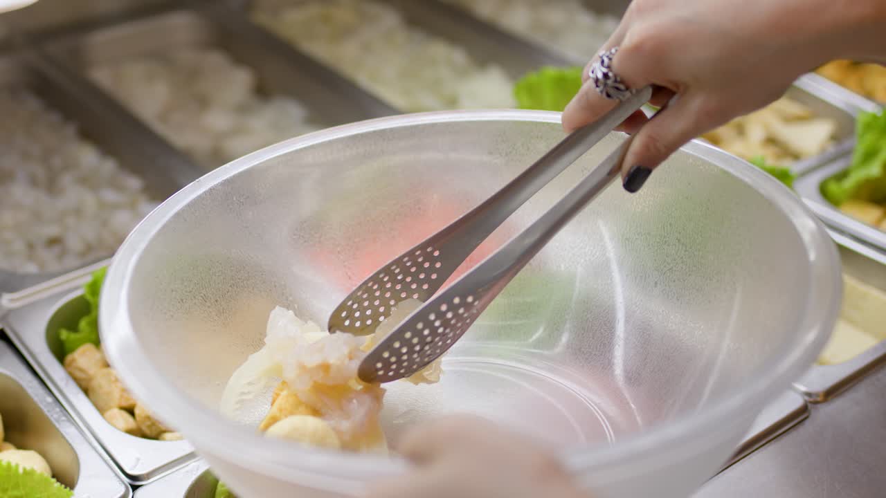 Hand uses tongs to pick seafood and vegetables into bowl at bright buffet food station