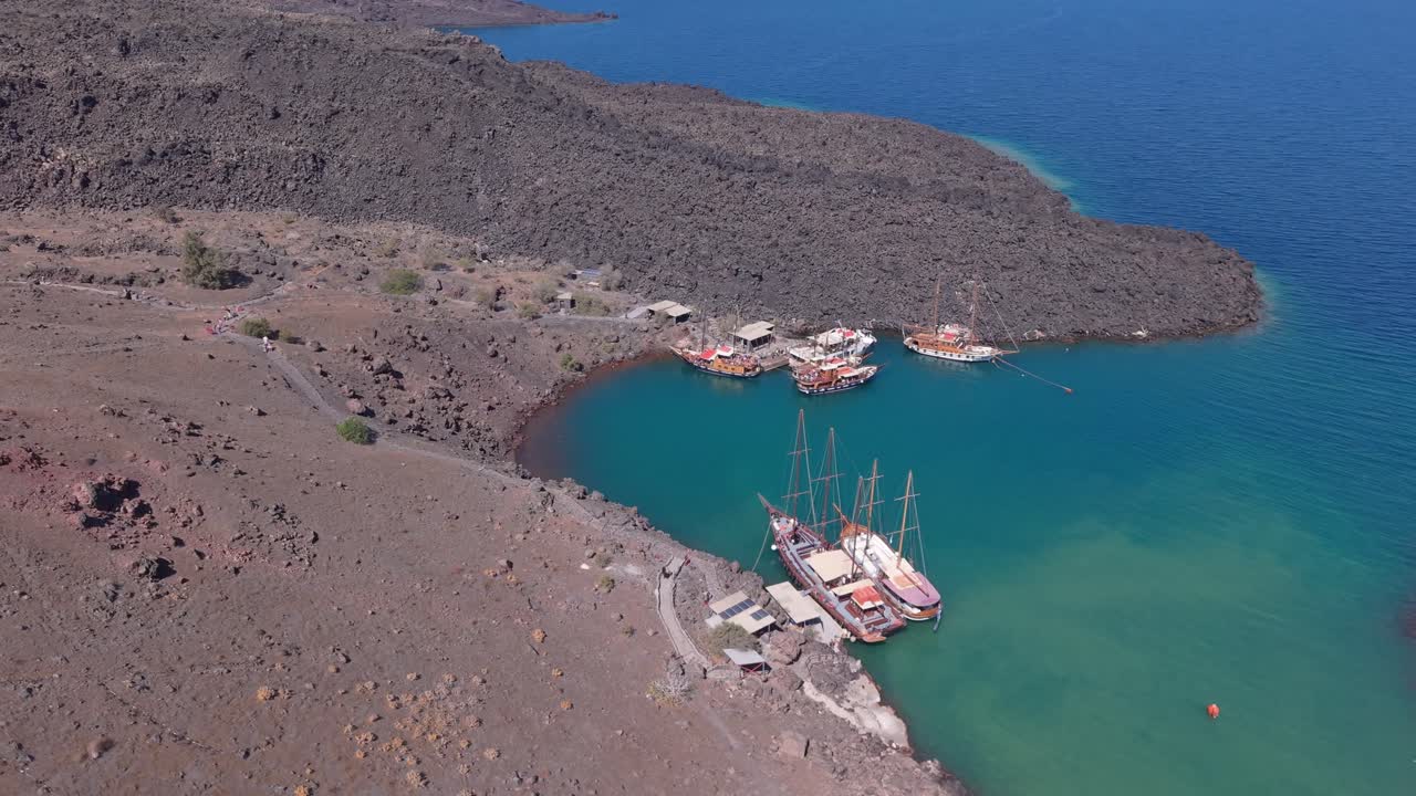 Drone orbiting the volcanic harbor of Santorini with a tourist boat, showcasing rugged terrain, caldera waters, and the dramatic Aegean landscape