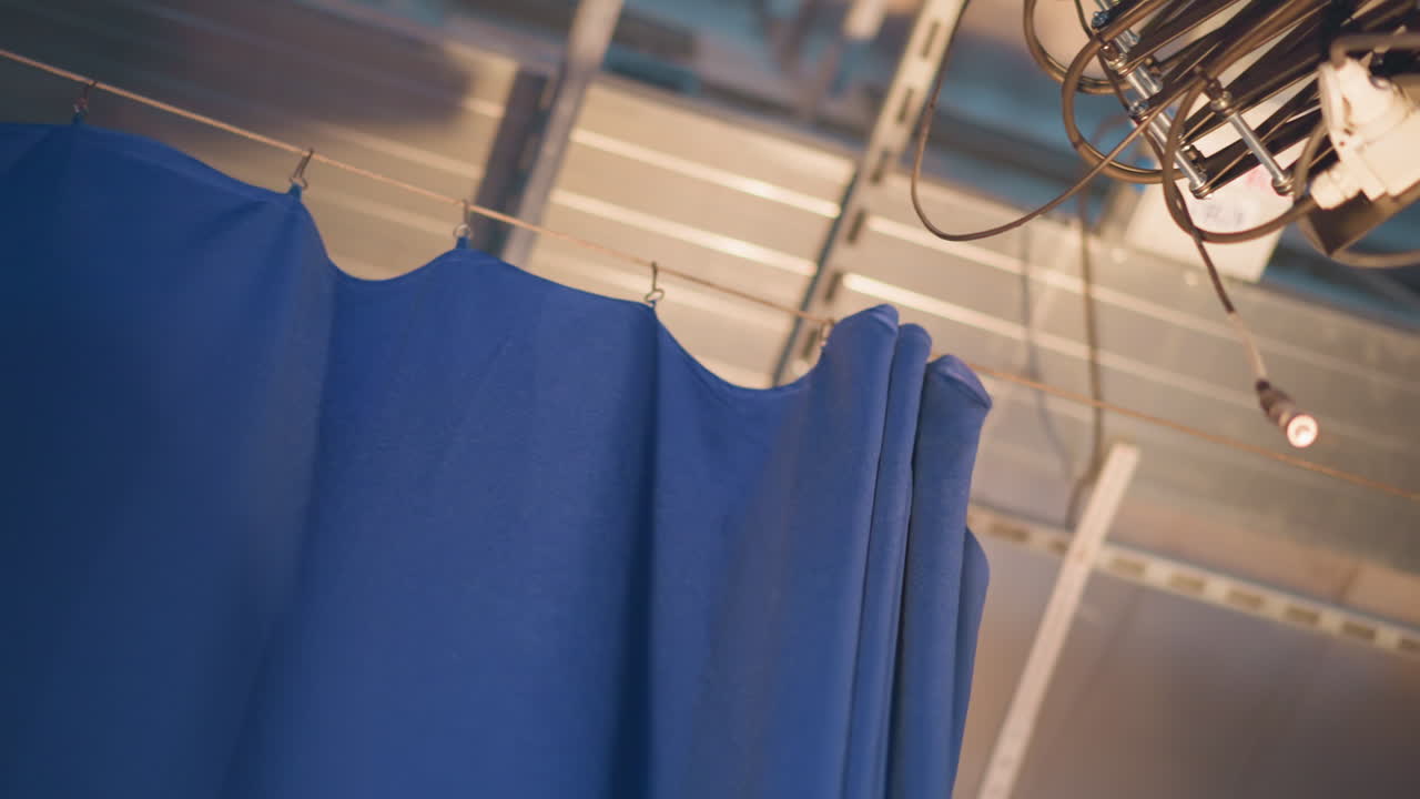 Overhead rigging suspends heavy blue curtain inside television studio, closeup view of metal clips and cables securing fabric to ceiling track, backstage environment highlighting production equipment and set design