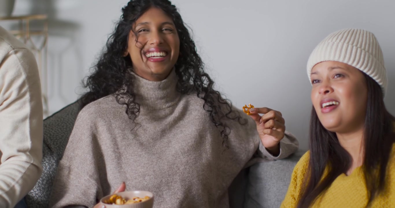 Amigos afro-americanos conversando no sofá, mulher central segurando um pretzel, rindo enquanto um homem entra.