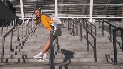 attractive cute young hipster millenial woman student having fun riding a banister and listening to music at urban background. wearing backpack, yellow blouse and silver skirt