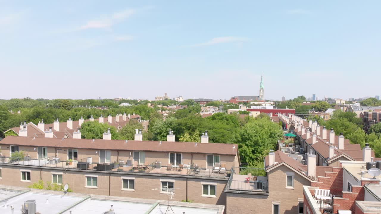 Wide aerial shot of a row of brick and concrete buildings with flat roofs, captured as a drone ascends to reveal a cityscape under a clear blue sky