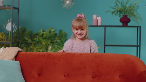 Child-girl-playing-hide-and-seek-peekaboo-game-near-sofa-at-home-alone-looking-at-camera-and-smiling