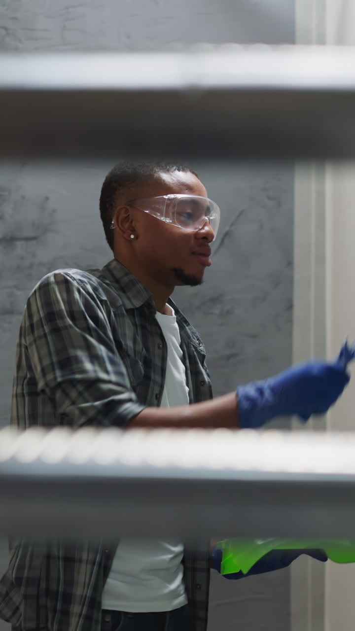 Positive black man paints wall with blue color in living room view from under step ladder. African-American worker renovation of apartment interior