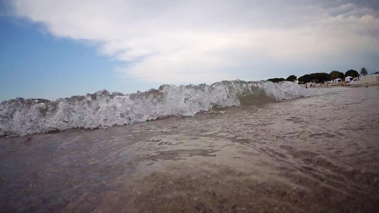 Primo piano dal basso di onde marine che si infrangono su una spiaggia sabbiosa sotto un cielo nuvoloso e drammatico, con palme e lettini in lontananza.
