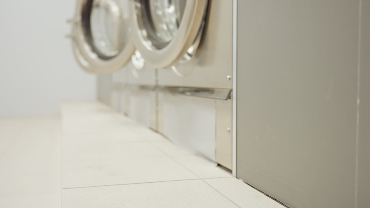 Low angle closeup hand placing black bucket on tiled laundromat floor beside stainless front load washing machines, beginning cleaning routine inside bright utility room, focus on equipment