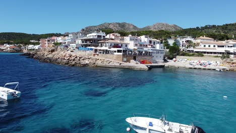 Drone-Alejándose-Hacia-Atrás-De-Casas-Y-Edificios-Portuarios-Sobre-Aguas-Claras-Y-Barcos-En-Un-Puerto-Natural-En-Mallorca-España