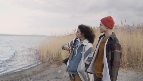 Side-View-Of-A-Teenage-Boys-Talking-Standing-Near-Of-Seashore-On-A-Cloudy-Day,-Other-Friend-Is-Crouched-Taking-Things-Out-Of-A-Backpack