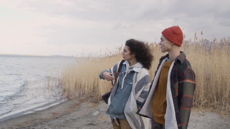 side view of a teenage boys talking standing near of seashore on a cloudy day, other friend is crouched taking things out of a backpack