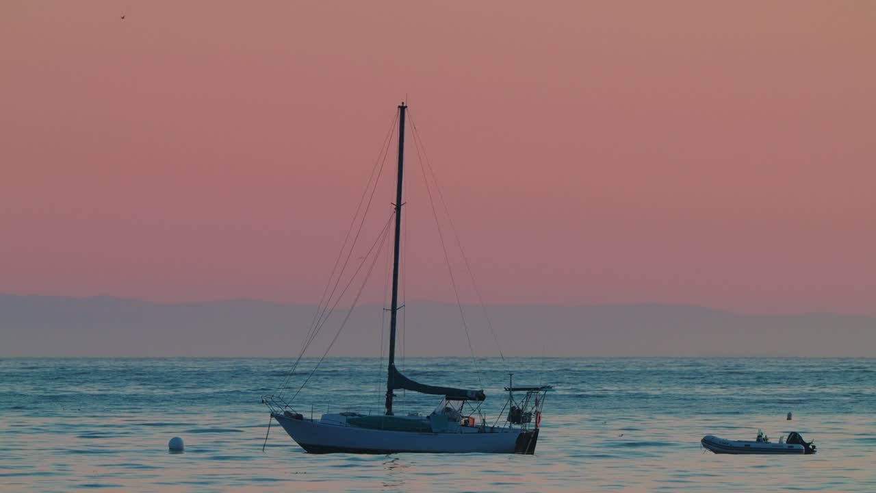 Lone sailboat drifts peacefully on calm waters, bathed in soft sunset hues, creating a tranquil maritime scene.
