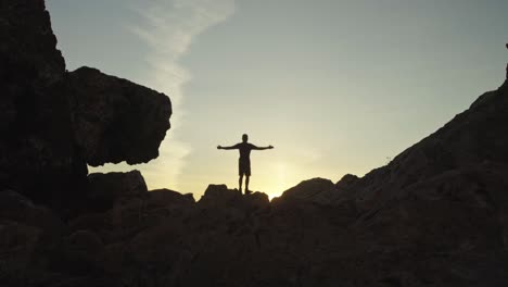 Un-Hombre-Levanta-Los-Brazos-Al-Atardecer-Rodeado-De-Una-Estructura-épica-De-Acantilados