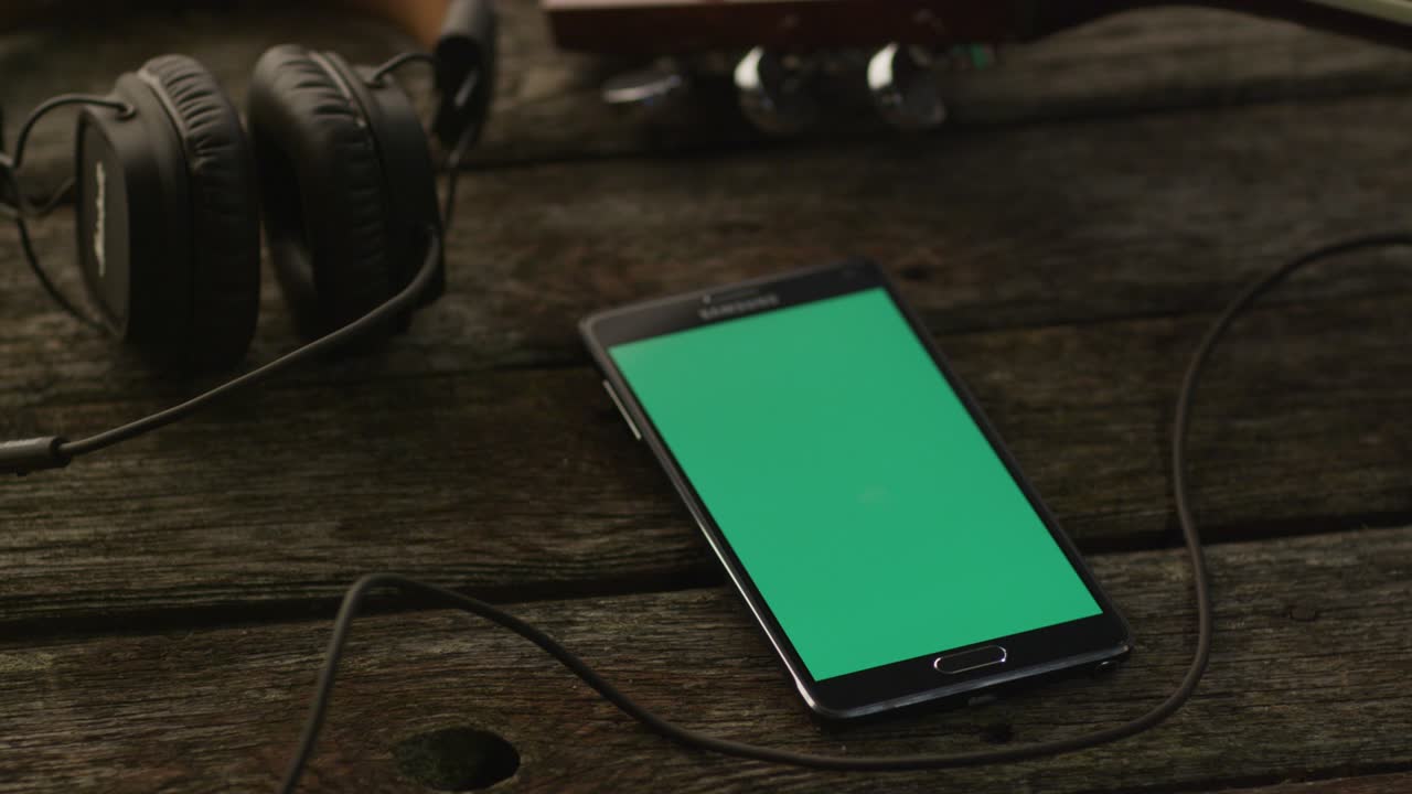 초록색 화면을 가진 안드로이드 드폰은 포트레이트 모드에서 기타와 헤드폰 의 나무 테이블에 놓여 있습니다.