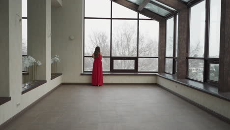 mujer en vestido rojo mira por la ventana de invierno. joven modelo femenino complementa las ventanas panorámicas de la galería con presencia graciosa. encanto y elegancia