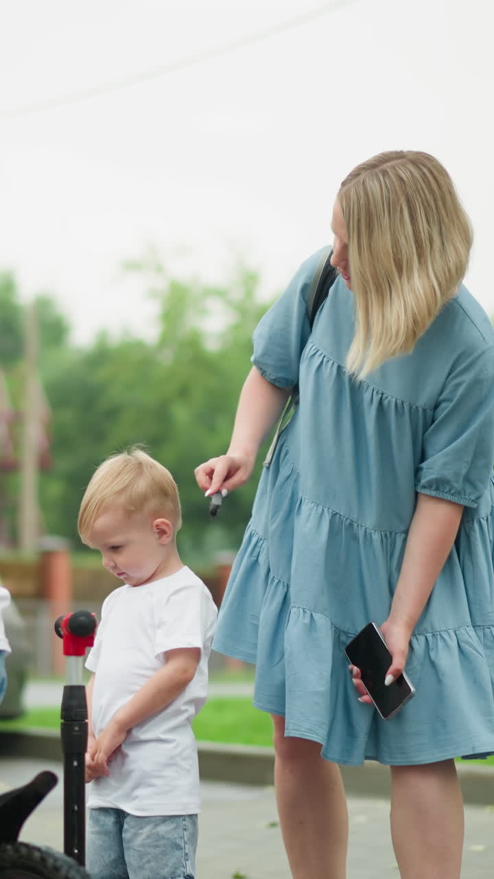 어머니는 조용하고 사려 깊게 보이는 어린 아들의 머리카락을 는 동안 구부러집니다. 큰 형제는 자전거를 들고 에 서 있고, 빨간 스쿠터는 근처에 있습니다.