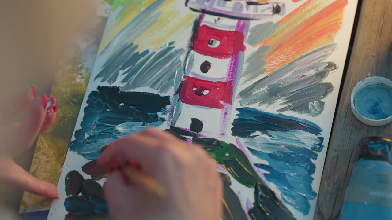 high angle view of artisan hand applying wide brush strokes to background of vibrant painted tower on paper outdoors under warm sunset light capturing rich texture and color depth