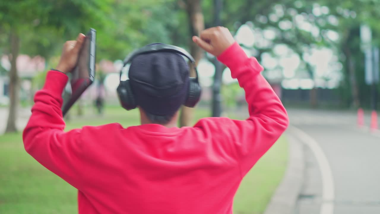 Rear View Of Young Man Walking On Sidewalk, Raising Hands Dancing While Listening Music From Wireless Headphones