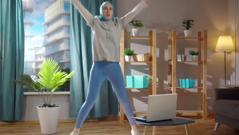 young muslim woman plays sports at home in the living room in front of her laptop