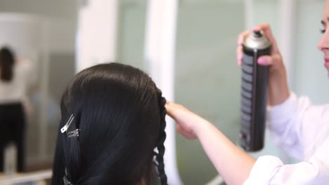Female-stylist-hands-applying-hairspray-fixing-plaiting-hairstyle-on-brunette-hair-back-view-slow-motion.-Hairdresser-finishing-hairdo-making-final-touch-with-spray