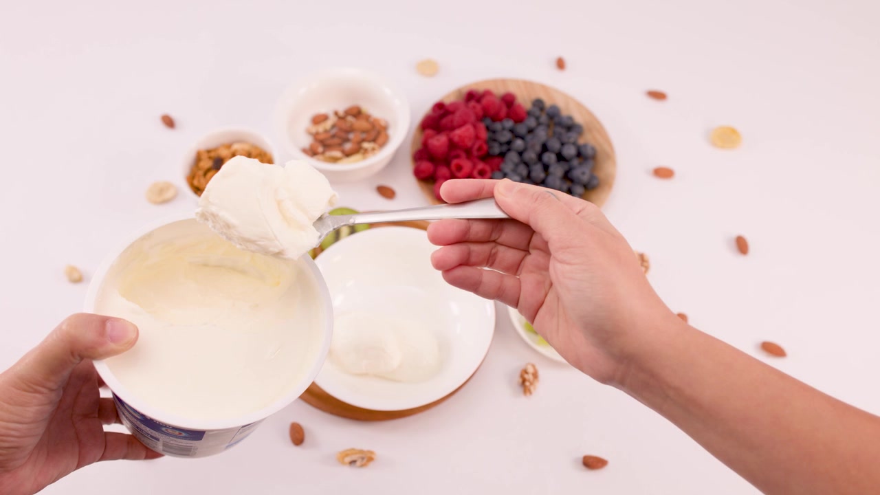 Kädet kauhovat kermaista jogurttia kulhoon, jota ympäröivät marjat, kiivi, granola ja mantelit.