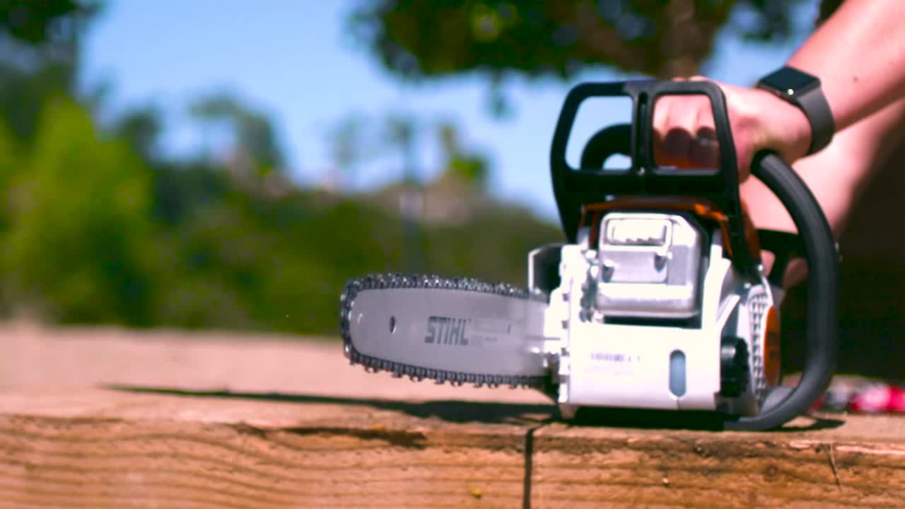 Watermelon being cut in half by a chainsaw in slow motion during summer time while sun is shining. Watermelon is focus and the background is slightly blurry in a cinematic way. Trees and table visible