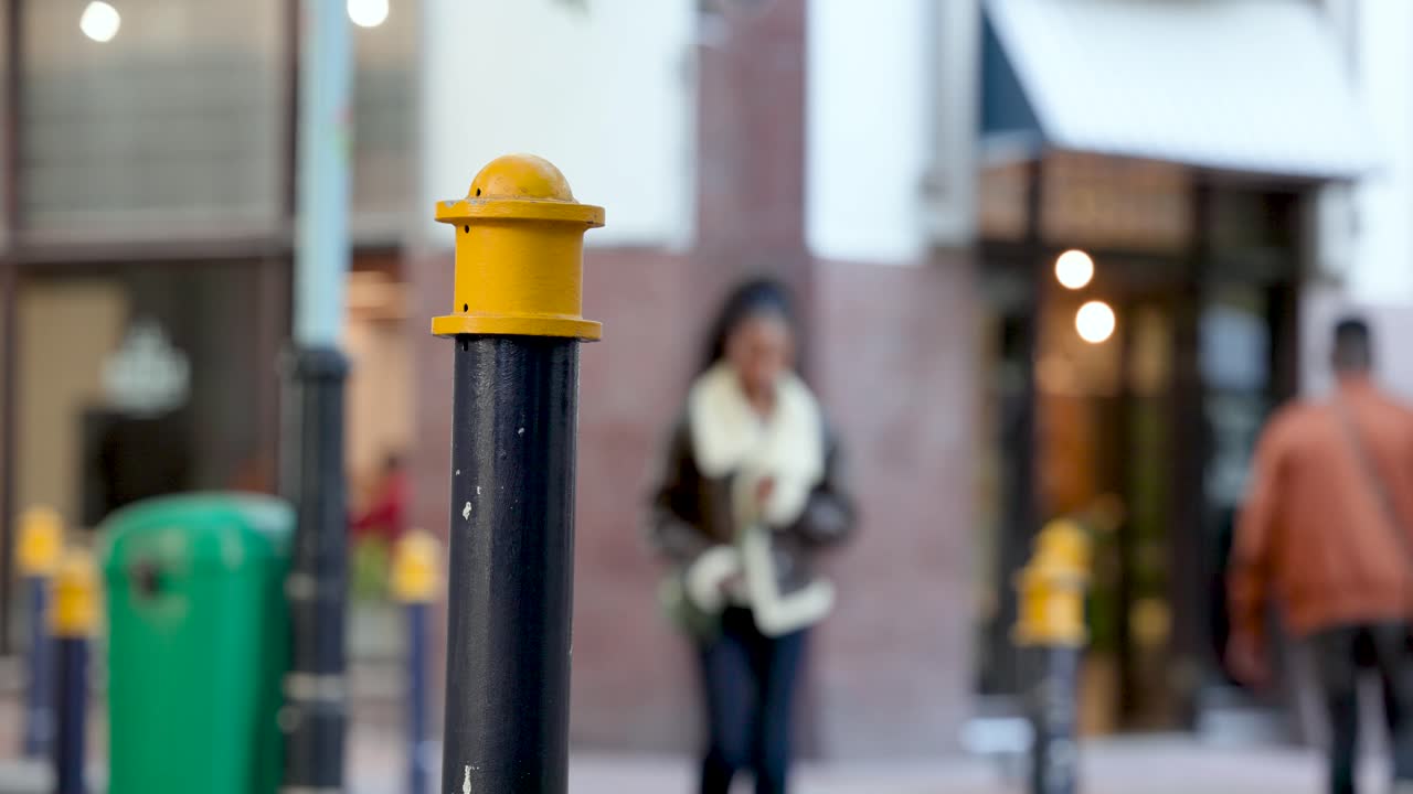 Forskellige fodgængere begynder at gå til butikkerne, mand i brun jakke passerer pullert med gul top