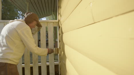 female painter wearing mosquito head net paints exterior wooden wall yellow