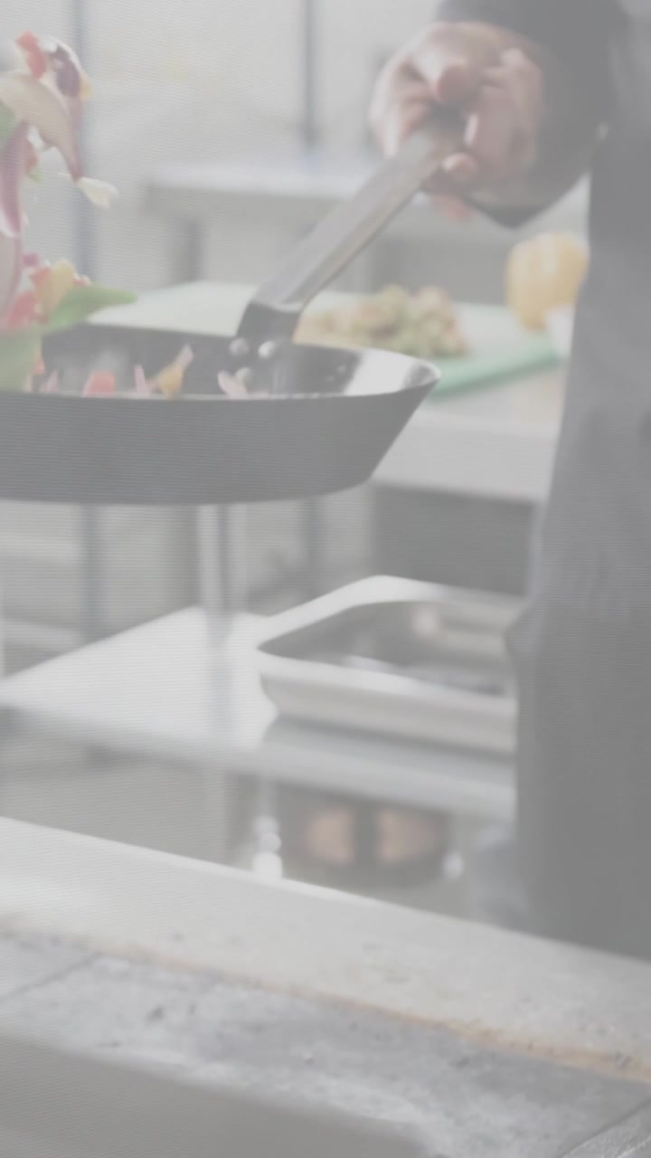 Video verticale: Chef che salta le verdure per il servizio in una cucina affollata, con la padella che si inclina e le pinze che guidano.