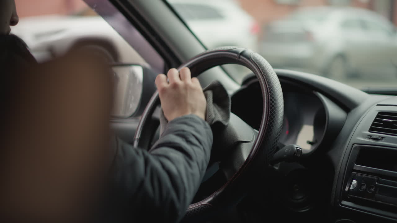 Back view of driver in car with window down wiping leather steering wheel in circular motion with towel focusing on driver expression closeup showing careful interior maintenance routine