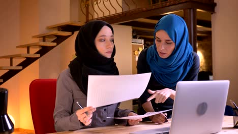 closeup portrait of two successful muslim businesswomen in hijabs comparing the data on different graphs using the laptop indoors in the office