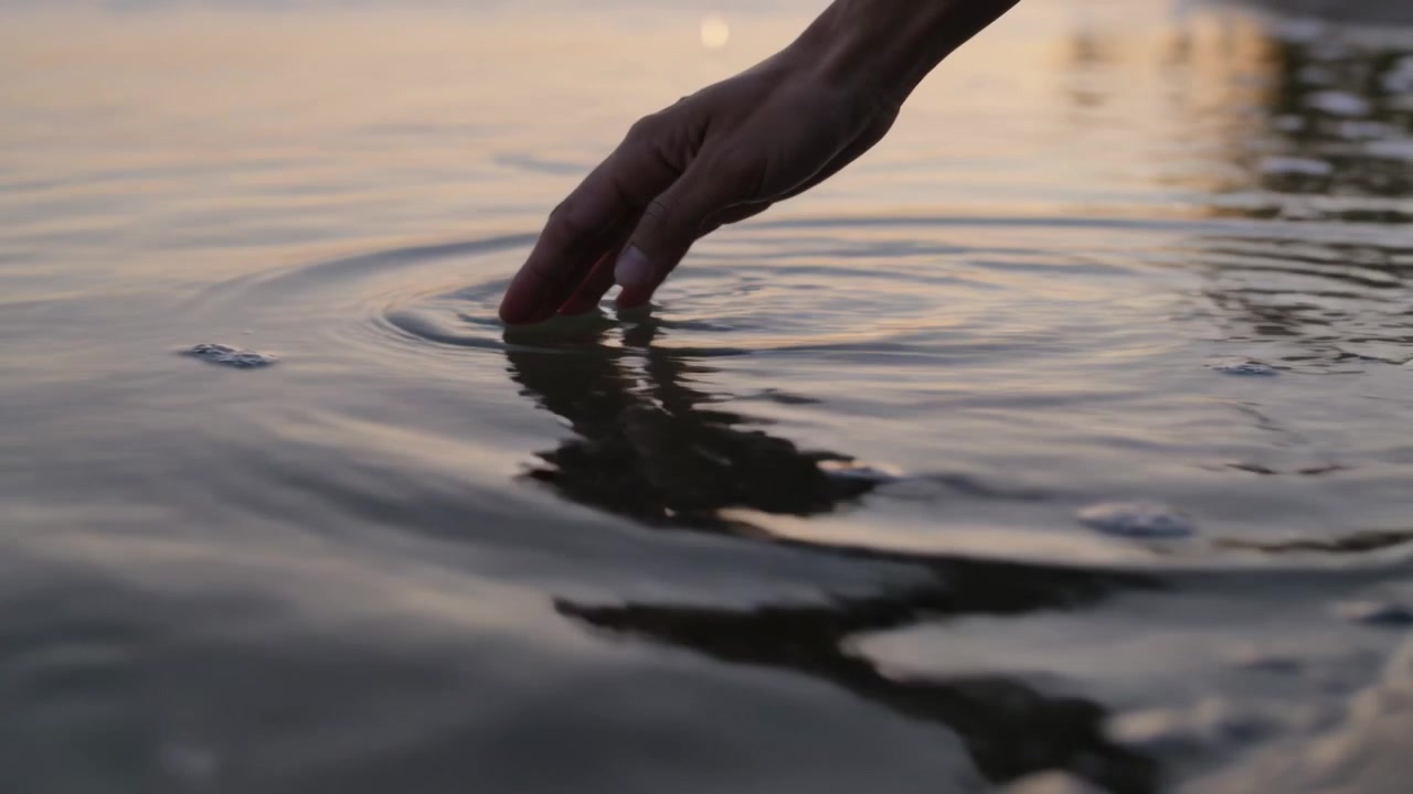Mano destra sospesa che crea increspature, abbassandosi per toccare l'acqua della battigia, percependo il riflesso, con spazio per il testo.