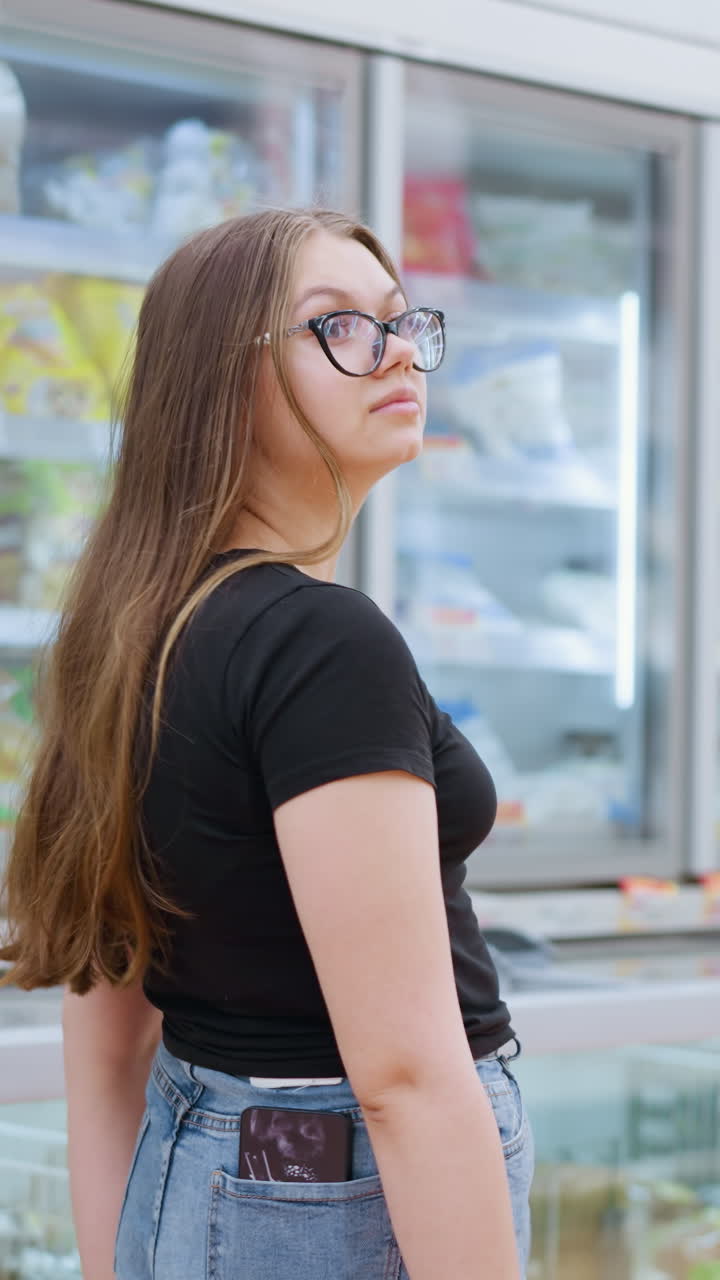 검은 과 안경을 입은 여자가 잘 조명 된 쇼핑몰에서 상품으로 가득 찬 전시 유리창을 지나가는 동안 오른쪽을 쳐다보고, 그녀는 집중하고 주변에 참여하는 것처럼 보입니다.