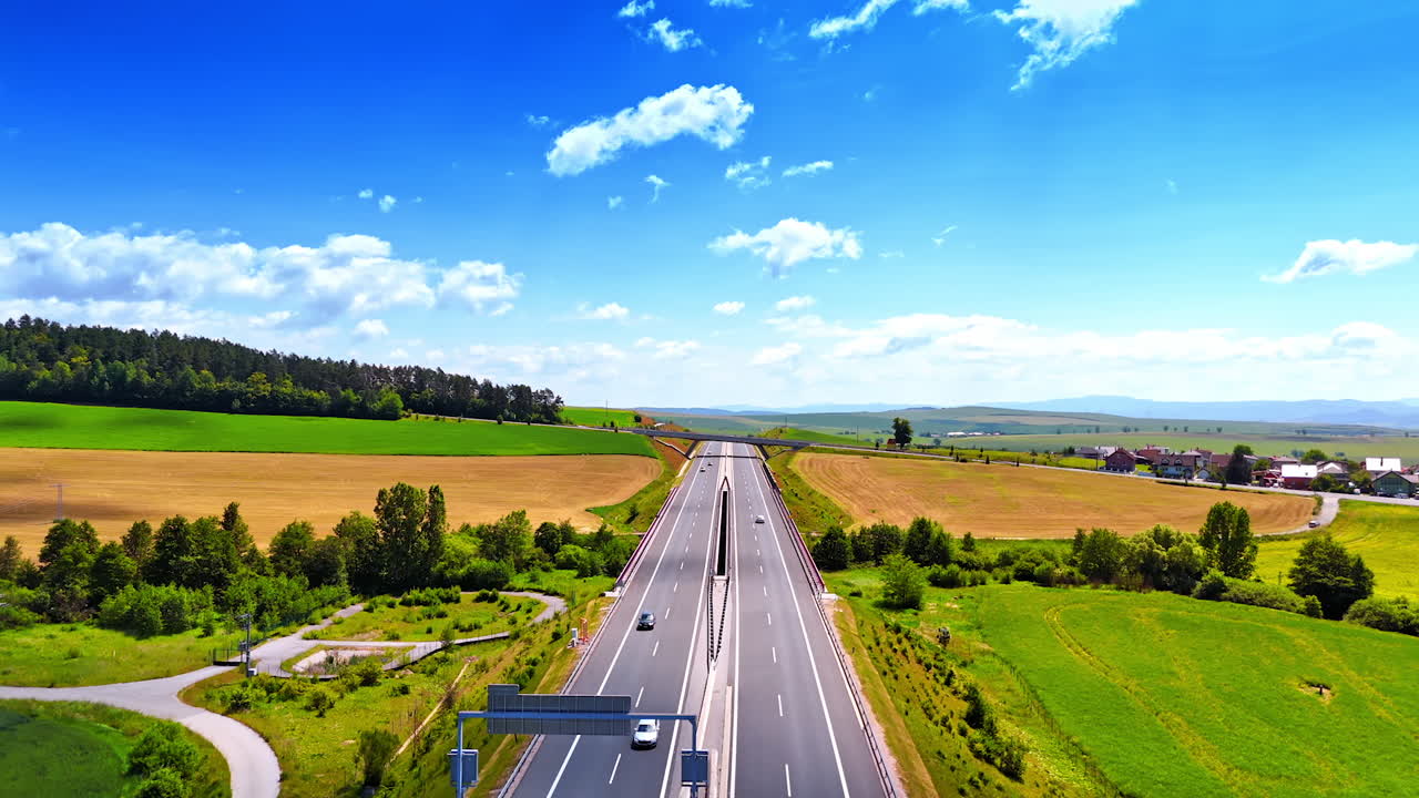 Daylight drive through green fields. A highway stretches through lush green fields under a clear blue sky with few clouds. Cars travel along the road