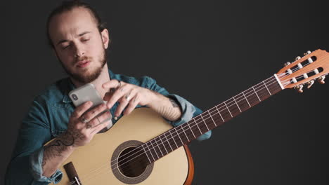 joven caucásico revisando su teléfono inteligente mientras sostiene la guitarra.