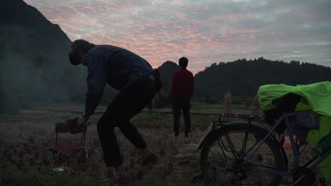 captivating sunset scene in vietnam's hajan loop with a silhouetted figure, field, and cycle near a warm campfire, under a colorful evening sky, creating a tranquil mood