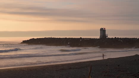 Silhouette-Eines-Nebelhorns-Und-Einer-Person,-Die-Wellen-Am-Strand-In-Bandon,-Oregon,-Bei-Sonnenuntergang-Beobachtet---Totale