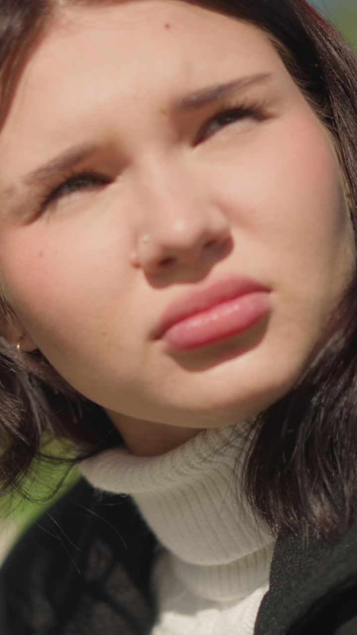 primer plano de una mujer al aire libre mirando a la luz del sol, con un abrigo negro y un suéter blanco, sosteniendo un teléfono, expresión reflexiva con un fondo borroso de vegetación y árboles