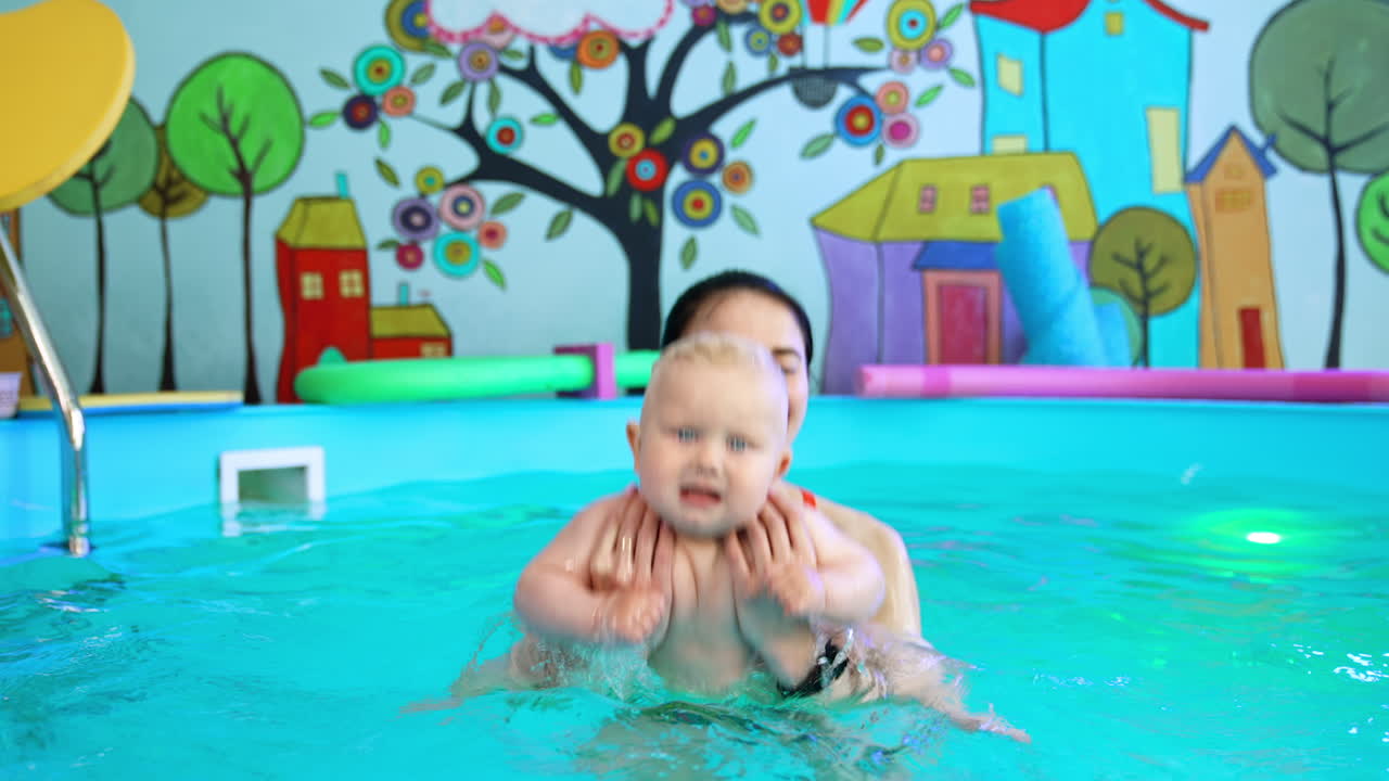 Female coach holding a lovely blond baby dipping him in the water. Infant is taught to swim in the swimming pool.